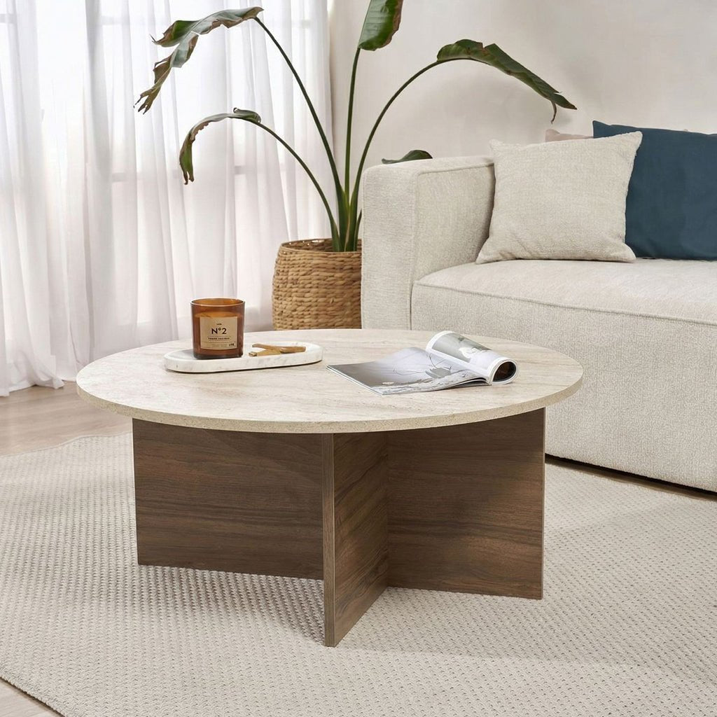 Mesa de centro redonda con tapa de travertino y base de madera oscura en forma de cruz. Banco y planta al fondo.