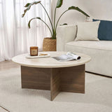 Mesa de centro redonda con tapa de travertino y base de madera oscura en forma de cruz. Banco y planta al fondo.
